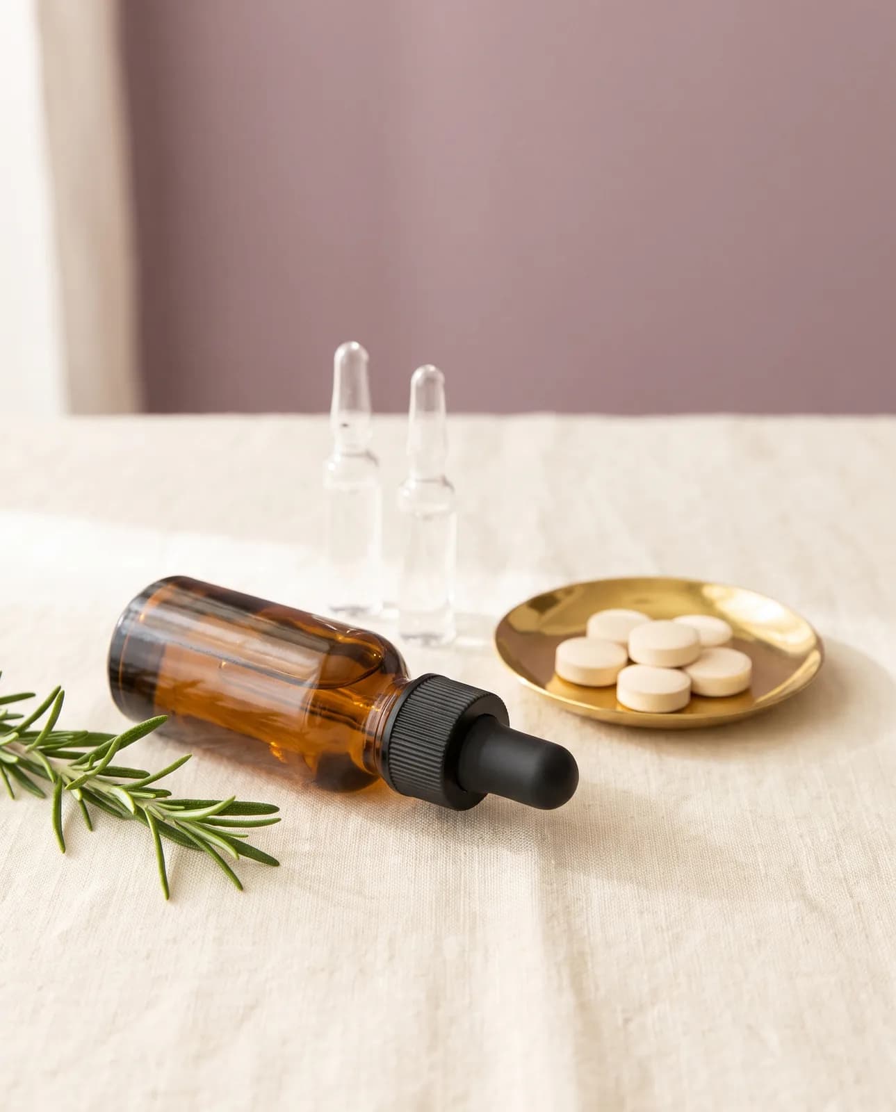 A premium vanity tableau for hair, lash, and skin care — plain tablets in a brass dish, an unlabeled amber dropper bottle, and clear glass ampoules on cream linen in soft morning light